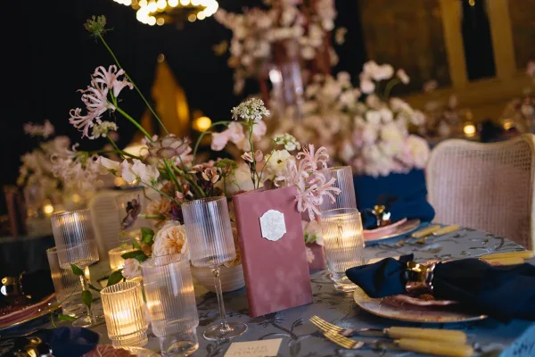 Reception tablescape in a dusty rose and navy table setting, centered by a blush floral centerpiece beneath warm chandelier lights.
