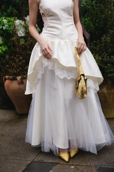 Wedding dress close-up of an ivory satin bridal gown with a yellow patterned umbrella amid garden greenery and wet terracotta planters.