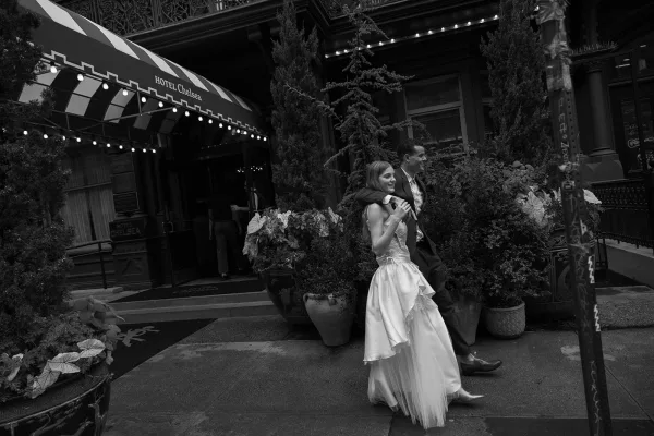 Bride and groom walking arm in arm for a couple portrait under a striped hotel awning, city sidewalk framed by potted plants and string lights.