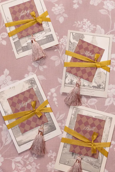 Wedding escort cards styled as tassel place cards, neatly arranged on pink floral fabric over a linen tablecloth.