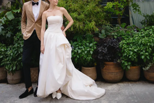 Couple portrait of a bride and groom standing, her ivory strapless ballgown with a train on a patio bordered by potted greenery.