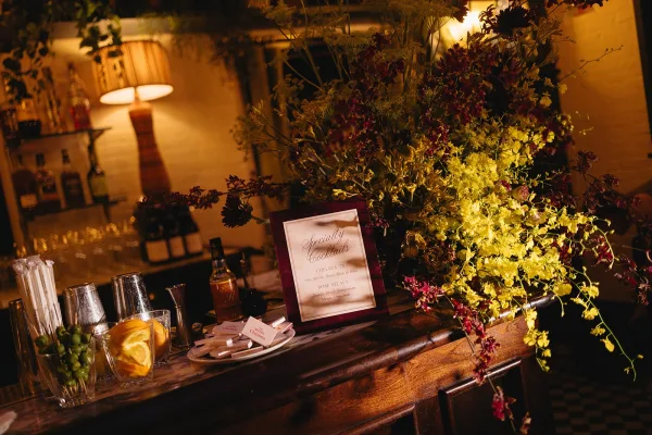 Signature cocktail sign anchors the wedding bar decor, paired with florals on a wooden bar beneath hanging greenery and a brick wall.