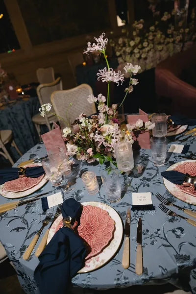 Reception tablescape on a blue floral tablecloth, candlelit and flanked by cane chairs in a dim interior.