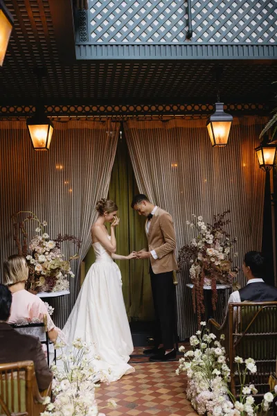 Ring exchange at an indoor ceremony as the groom places the band and the bride wipes happy tears, under hanging lanterns with striped curtains behind.
