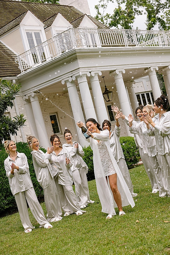 Bridesmaids champagne pop as the bridal party in pajamas cheers in white satin sets, champagne spray flying outside a columned mansion