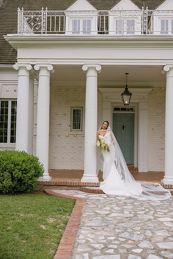 Bridal portrait of a bride holding bouquet in a strapless satin gown with cathedral veil, leaning on a white columned porch with lantern backdrop