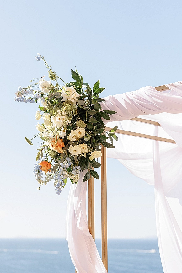 Wedding ceremony arch with asymmetrical arch flowers, sheer draping, and pastel blooms on a wooden frame set against the ocean horizon