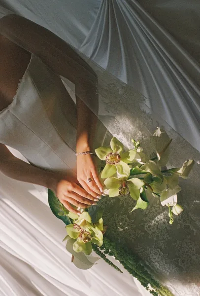 Bridal bouquet of green orchid bouquet with white blooms, held in manicured hands with wedding ring and bracelet against soft draping