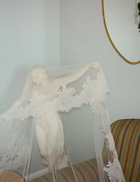 Bridal veil portrait of a bride draped in a lace cathedral veil over her face, wearing a strapless satin dress against a white wall mirror backdrop