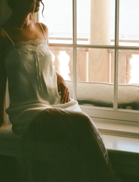 Bridal portrait of a bride by window in a lace-trim slip, showing her engagement ring with an ocean-view balcony behind