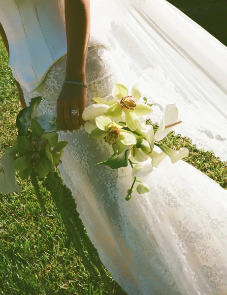 Bridal bouquet with orchid bridal bouquet and calla lilies, held by a bride in lace dress and veil on a sunlit grass lawn