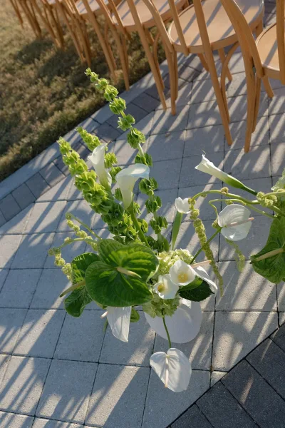 Wedding aisle flowers in white vase with calla lilies and anthurium, lining a stone paver walkway beside wooden chairs in garden greenery