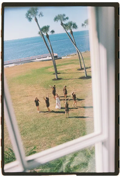 Bride with bridesmaids in black dresses cheering with arms up on an oceanfront lawn, her strapless gown train flowing by palms