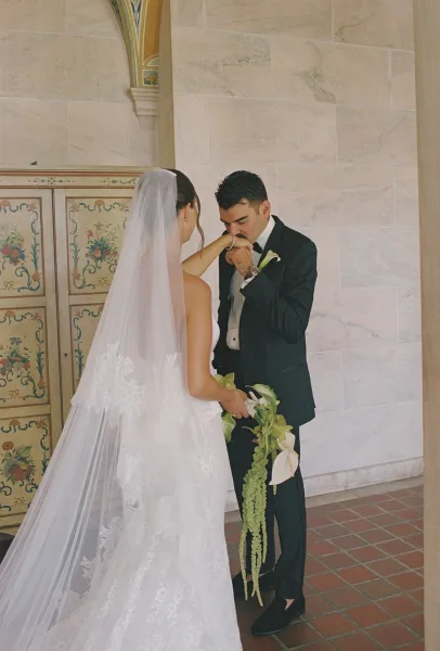 Wedding first look as the groom kissing bride hand while they hold hands, her lace veil and calla lily bouquet by a stone wall doorway