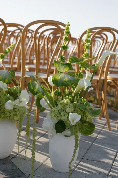 Ceremony aisle decor with aisle floral arrangements in white ceramic vases, calla lilies and greenery beside wooden chairs on patio pavers