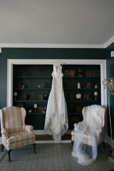 Wedding dress with lace wedding dress details on a hanger, veil draped beside it in a vintage library with bookshelves and lamps