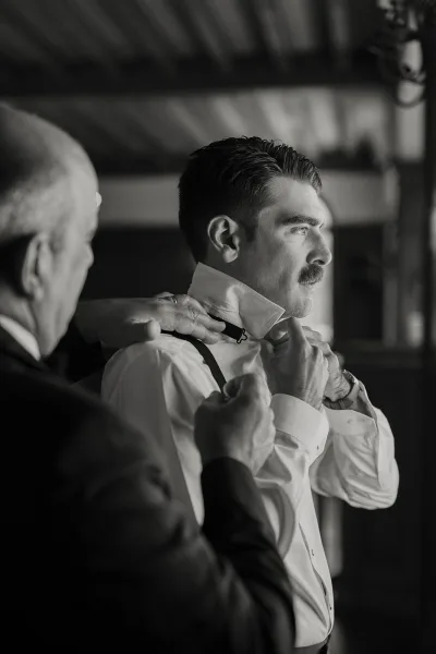 Groom getting ready as his father adjusts his bow tie, suspenders and wristwatch in soft window light beneath ceiling beams