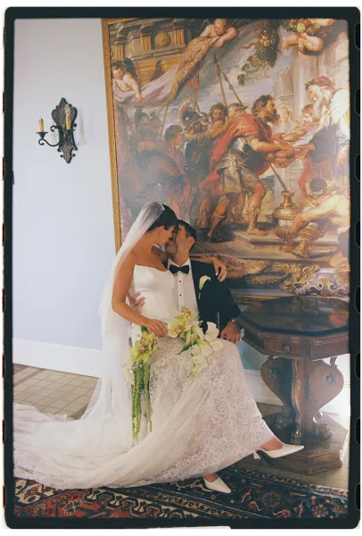Wedding kiss portrait of bride sitting on groom in a strapless gown and veil, holding cascading orchid bouquet beside a framed mural indoors