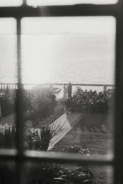 Wedding ceremony on a sunlit oceanfront lawn with an aisle runner, chair rows, and floral arrangements facing the shoreline horizon