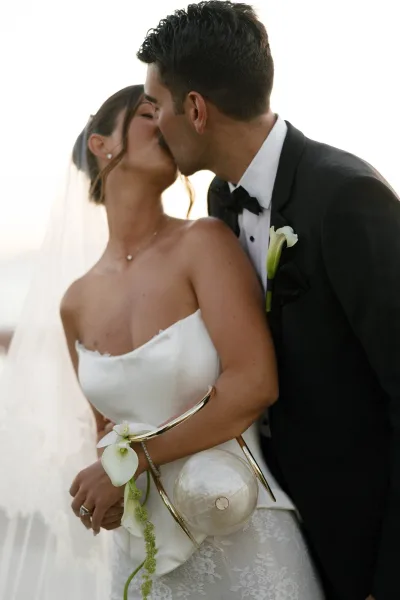 Wedding kiss portrait of bride and groom kissing, her veil and calla lily bouquet against a bright sky at an outdoor waterfront