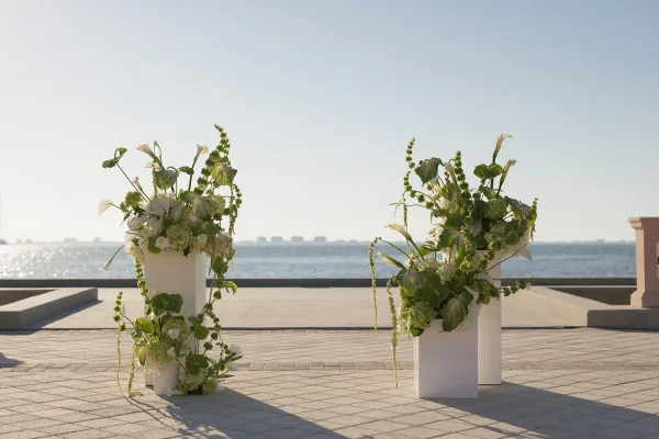 Ceremony floral pillars with white pedestal plinth flowers and greenery on a paved waterfront terrace overlooking the ocean horizon