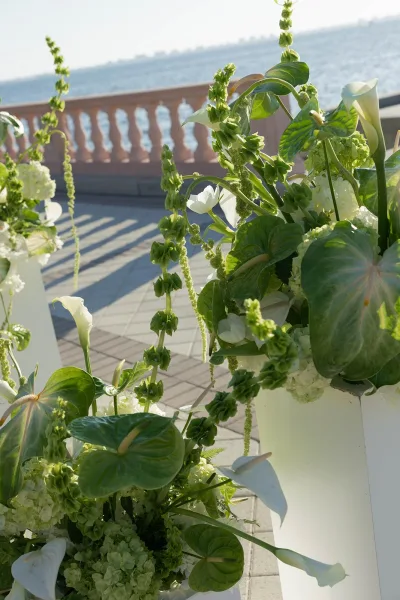 Wedding aisle florals in white calla lilies and hydrangeas cascade over a pillar with greenery on a tiled patio by the waterfront