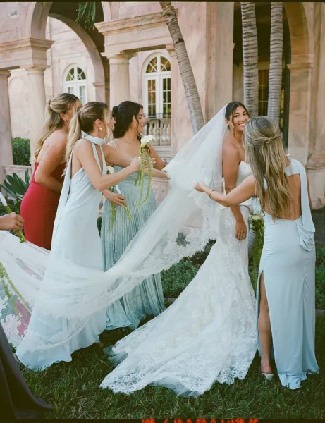 Bride with bridesmaids adjusting her long lace wedding veil and train as she looks back on a lawn by a stucco building with palms