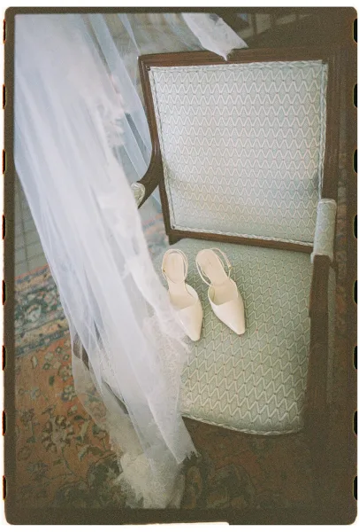 Bridal shoes, white wedding heels with pointed toes on an upholstered chair, a tulle veil draped over lace trim beside them