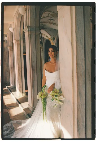 Bridal portrait of a bride holding bouquet of orchids and calla lilies in a strapless lace gown, posed in a sunlit stone archway corridor