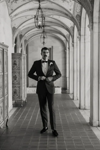 Groom portrait in a black tuxedo, bow tie, and boutonniere, standing in an arched stone corridor with hanging lanterns overhead