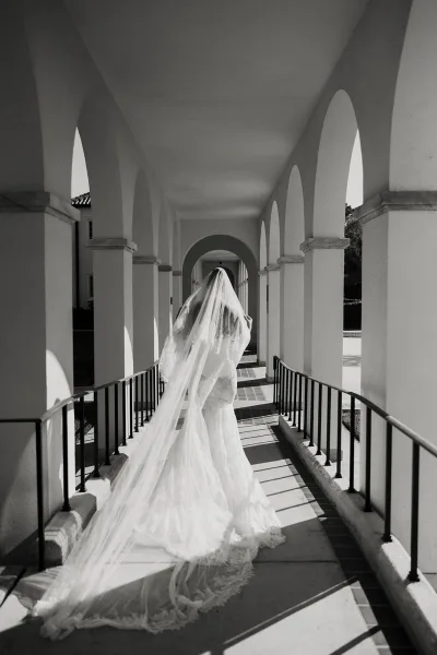 Bridal portrait of a bride from behind in a lace gown with a long veil train, walking through a sunlit arched colonnade corridor