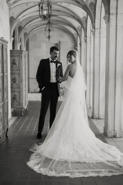 Couple portrait of bride and groom holding hands, her cathedral veil and lace train flowing in an arched stone hallway with lanterns
