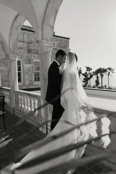 Wedding couple portrait of bride and groom touching foreheads, veil flowing over lace dress by an arched colonnade with ocean view