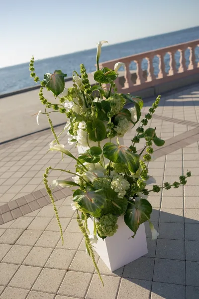 Wedding floral arrangement of white calla lilies and greenery on a pedestal, with ocean and sky beyond a stone terrace railing