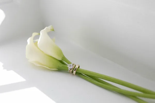 Wedding rings with engagement ring detail, diamond solitaire and gold bands resting on a calla lily in soft natural light on white surface