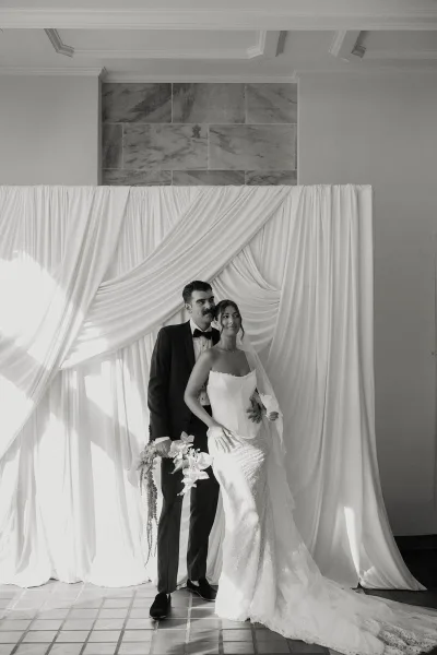 Couple portrait in a black and white wedding portrait style, groom in tuxedo behind bride in lace gown and veil holding orchid bouquet before draped backdrop