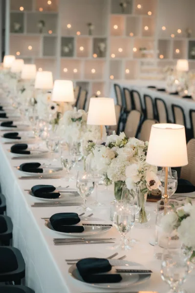 Reception tablescape on a long banquet table wedding with white floral centerpieces, table lamps, taper candles, and string-lit shelving backdrop