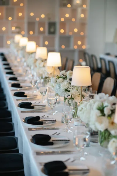Reception tablescape on a long banquet table with white linens, black napkins, floral centerpieces, lamps, and candles under warm string lights