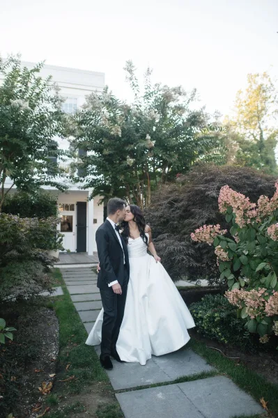 Wedding kiss portrait of bride and groom kiss on a garden path, bride in strapless ball gown with long veil beside greenery and white house exterior