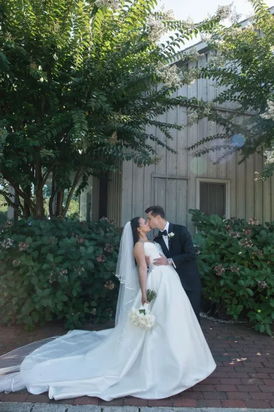 Wedding kiss portrait of bride and groom kiss, her cathedral veil and bouquet flowing as he dips her in sunlit garden by barn wall