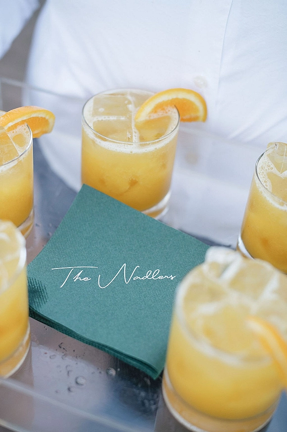 Wedding cocktail in rocks glasses with orange slices and ice on a silver tray, paired with teal napkins featuring white cursive lettering
