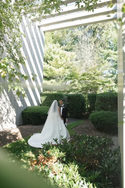 Couple portrait of bride and groom holding hands, her veil and dress train flowing beside his tuxedo on a sunlit garden walkway near a modern building