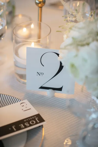 Wedding table number card displayed in a holder beside a votive candle, striped plate, and white floral centerpiece on a softly lit reception table