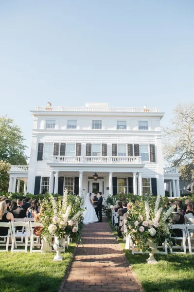 Outdoor wedding ceremony with a garden ceremony aisle of white chairs and hydrangea florals, bride and groom before a mansion facade under blue sky