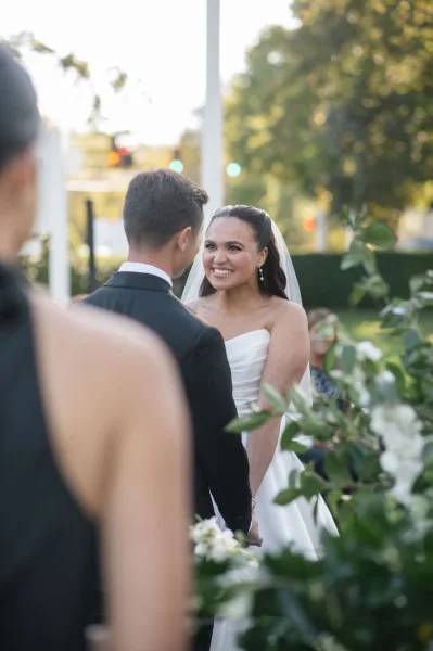 Ceremony moment as bride smiling at groom while holding hands, strapless gown and cathedral veil, white bouquet in a garden setting.