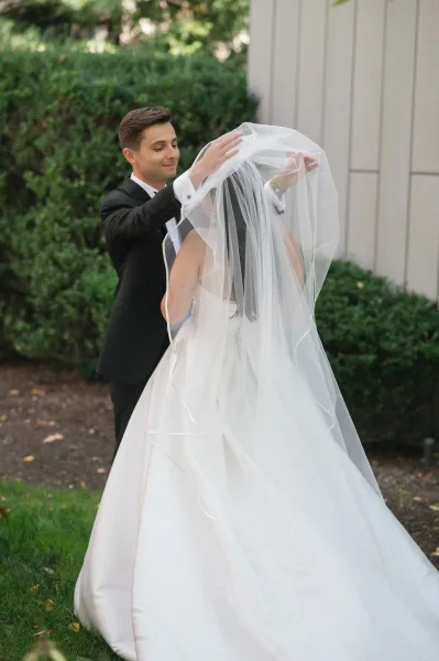 First look moment as groom lifting veil of bride in strapless ball gown, with long veil flowing by hedge greenery and gravel path