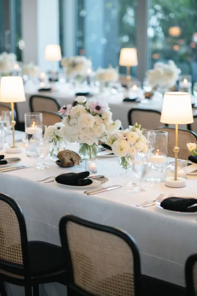 Reception tablescape with a white and black table setting, roses and hydrangeas in glass vases, votive candles and gold lamps by windows at dusk