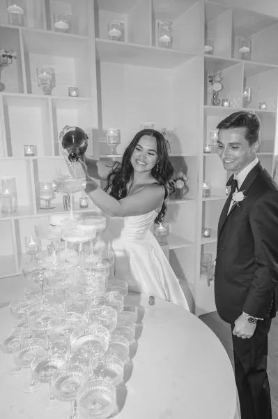 Champagne tower as bride in a wedding dress pours from a bottle while groom in tuxedo watches beside candles and flowers on white shelving wall