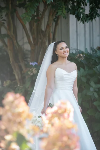 Bridal portrait of a smiling bride holding a white rose bouquet, wearing a long veil and strapless dress amid garden greenery and wood siding