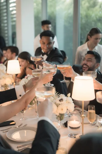 Wedding toast with champagne coupe glasses raised by tuxedoed guests at a candlelit table, window wall behind showing greenery outside
